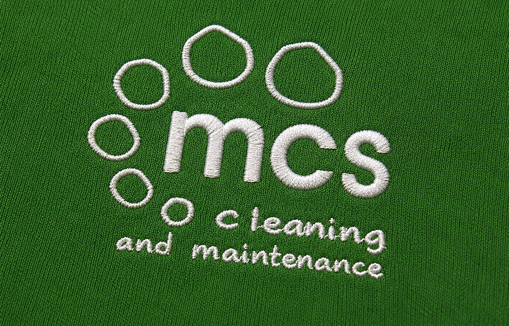 Close-up of the MCS Cleaning and Maintenance logo embroidered in white thread on a green fabric uniform