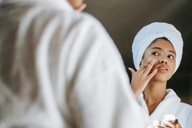 A woman with her hair wrapped in a towel applies skincare cream to her face while looking in a mirror.
