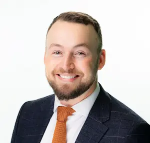 Professional headshot of a smiling bearded man wearing a navy blazer, white shirt, and orange tie against a white background