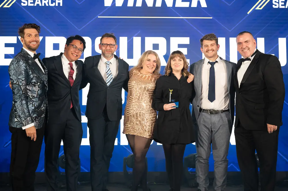 Formally dressed group posing on stage with an award trophy in front of a "Search Awards Winner" backdrop displaying "Exposure Ninja"