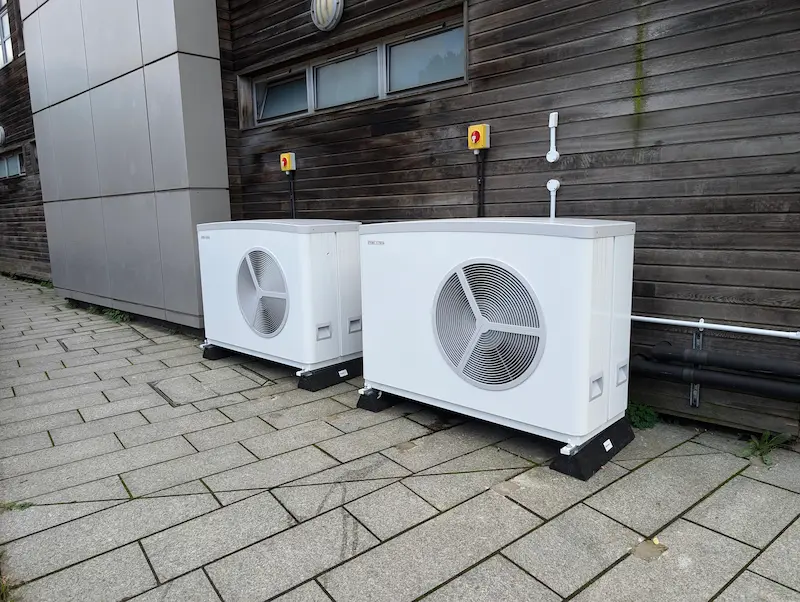 Two white air source heat pump units installed outside a building with wooden cladding, set on a paved stone walkway.