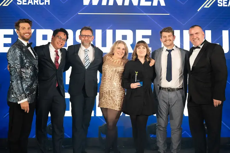 Group of seven people in formal attire posing on stage with a winner's trophy in front of a blue "Winner" backdrop at an awards ceremony