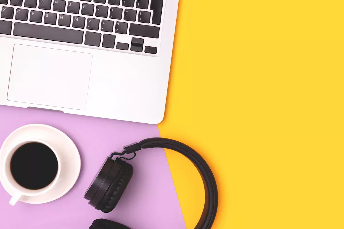 Overhead flat lay of a laptop keyboard, a cup of black coffee, and black headphones arranged on a two-tone purple and yellow background.