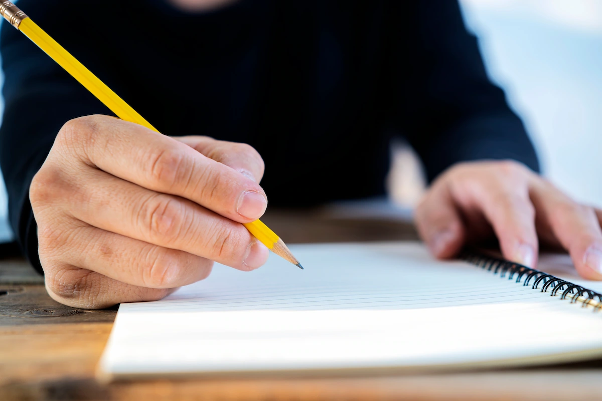 Close-up of a hand holding a yellow pencil about to write on a blank lined spiral notebook resting on a wooden table