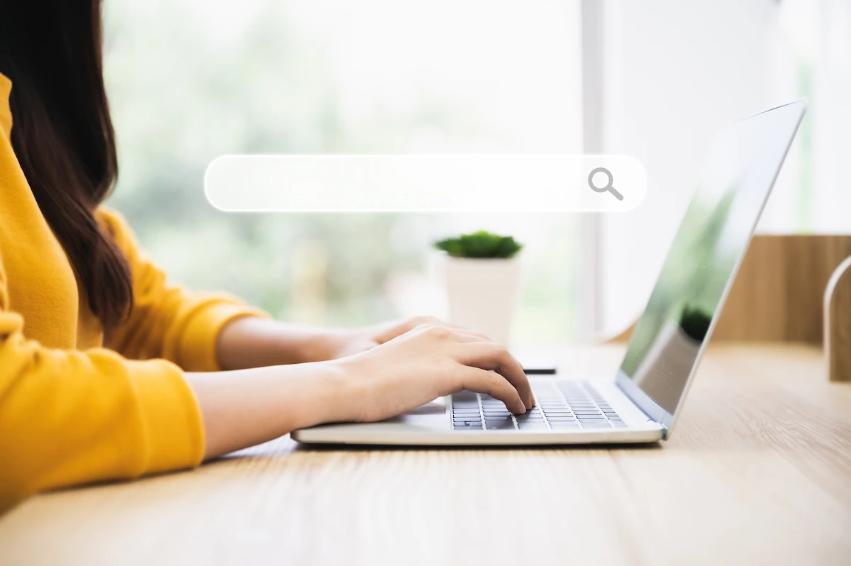 A woman in a yellow sweater types on a laptop, with a graphic overlay of an empty search bar floating above the desk.