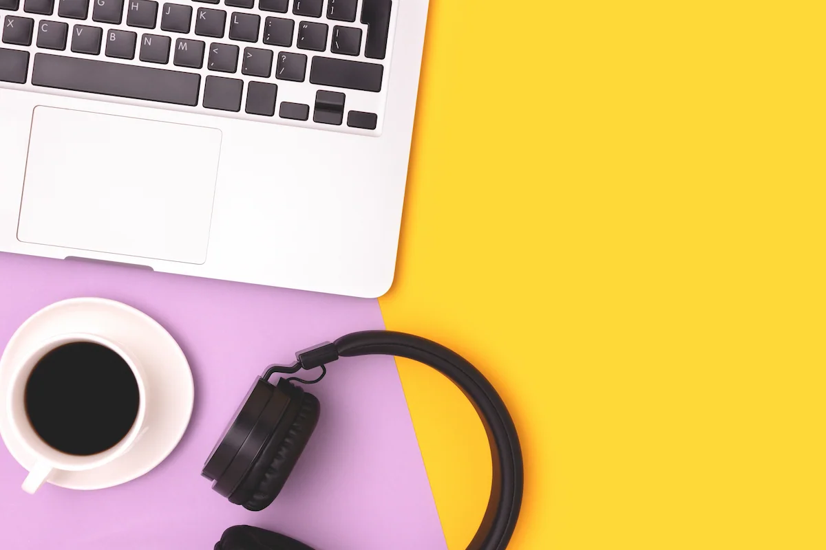 Overhead flat lay of a laptop keyboard, a cup of black coffee, and black headphones arranged on a two-tone purple and yellow background.