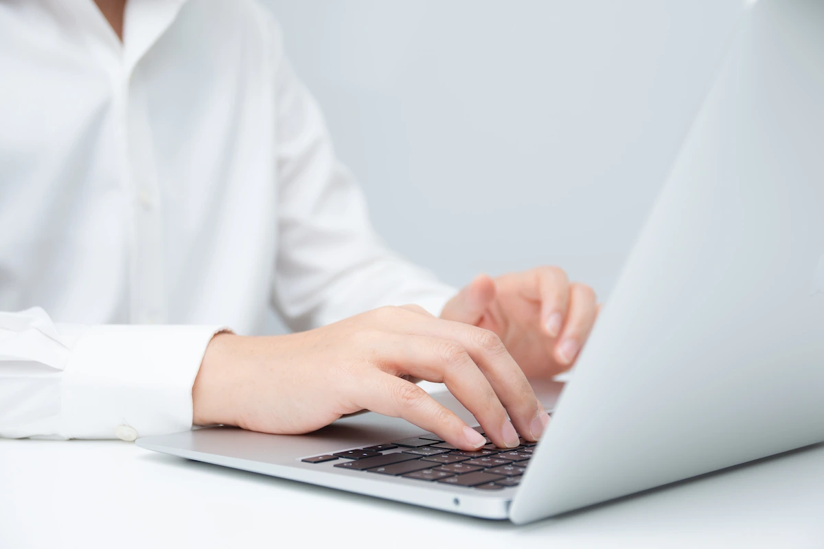 Close-up of a person in a white shirt typing on a silver laptop against a plain light gray background.