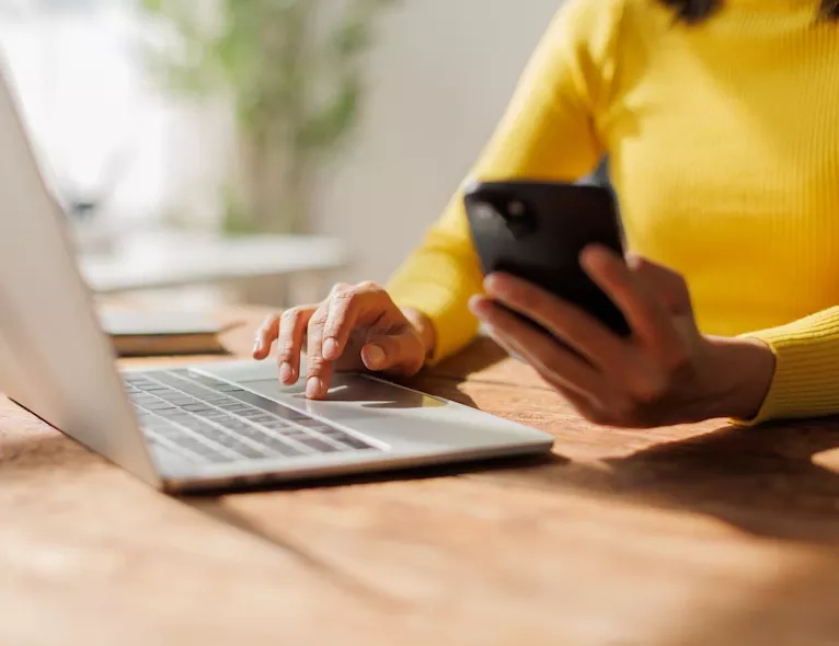 A woman in a yellow sweater uses a laptop trackpad with one hand while holding a smartphone in the other, seated at a wooden desk.