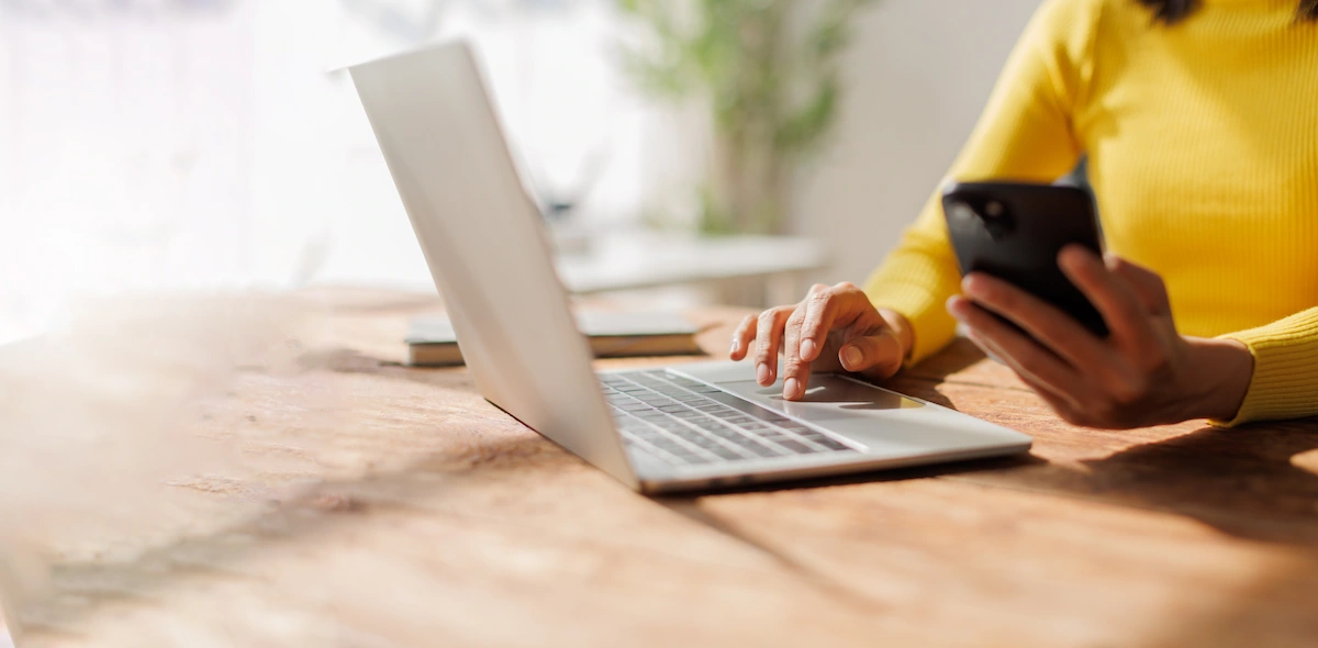 A woman in a yellow sweater uses a laptop trackpad with one hand while holding a smartphone in the other, seated at a wooden desk.